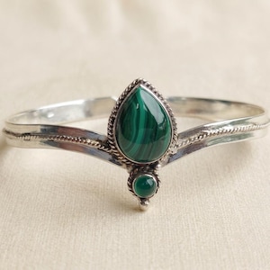 Puede incluir: Un brazalete de plata con una piedra de malaquita central en forma de lágrima con bandas verdes distintivas, acentuado por una piedra verde redonda más pequeña debajo. La banda de metal tiene una textura de cuerda trenzada en los bordes.