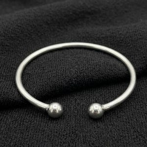 May include: A polished silver-tone cuff bracelet featuring a smooth, circular band that remains open at the front. The two ends of the bracelet are finished with small, spherical ball accents, creating a clean and simple design for the wrist.