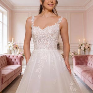 Puede incluir: Un vestido de novia blanco sin mangas con escote corazón y tirantes anchos. El corpiño y la falda están adornados con intrincados bordados de flores y hojas blancas que se extienden desde la parte superior hasta la falda de tul completa.
