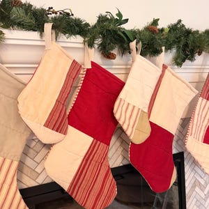 May include: A collection of quilted Christmas stockings featuring a patchwork design of solid red fabric, cream-colored fabric, and red-and-white striped fabric. Each stocking includes a fabric hanging loop at the top and horizontal stitching.
