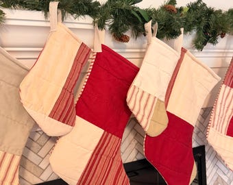 May include: A collection of quilted Christmas stockings featuring a patchwork design of solid red fabric, cream-colored fabric, and red-and-white striped fabric. Each stocking includes a fabric hanging loop at the top and horizontal stitching.