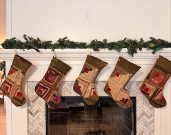 May include: Five Christmas stockings featuring a patchwork log cabin quilt design in shades of brown, tan, red, and cream. Each stocking has a solid olive green cuff at the top and is suspended by a white loop against a white fireplace mantel.