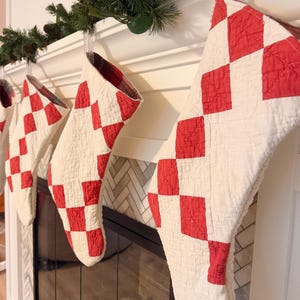 May include: Three quilted Christmas stockings featuring a red and white diamond pattern. Each stocking is constructed with a stitched fabric texture and includes a plaid fabric lining visible at the top opening, hanging from a white mantelpiece.