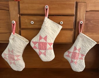 May include: Three white quilted Christmas stockings with pink star patterns hang from a wooden cabinet. Each stocking has a pink loop for hanging. The stockings are a festive holiday decoration.