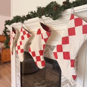 May include: Four quilted Christmas stockings hanging in a row, each featuring a red and white diamond checkerboard pattern. The stockings have a visible stitched texture and are lined with a red and black plaid fabric, suspended from a white mantel.