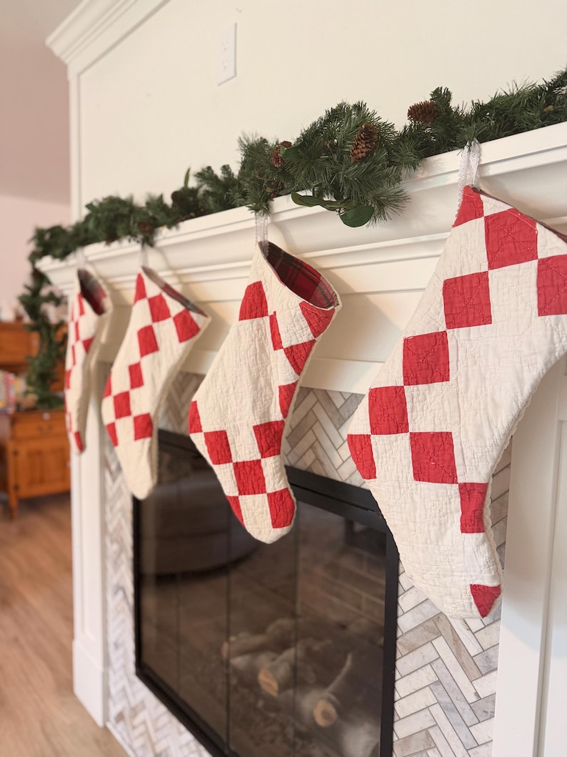May include: Four quilted Christmas stockings hanging in a row, each featuring a red and white diamond checkerboard pattern. The stockings have a visible stitched texture and are lined with a red and black plaid fabric, suspended from a white mantel.