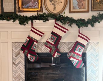 May include: Three quilted Christmas stockings hanging from a green pine garland. Each stocking features a patchwork design with red, dark green, and white fabric squares, accented by thin red and white striped bands at the top cuff.