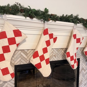 May include: Four quilted Christmas stockings featuring a repeating red and off-white diamond checkerboard pattern. Each stocking is constructed with visible stitching and hangs from a white mantelpiece, displaying a consistent geometric design.