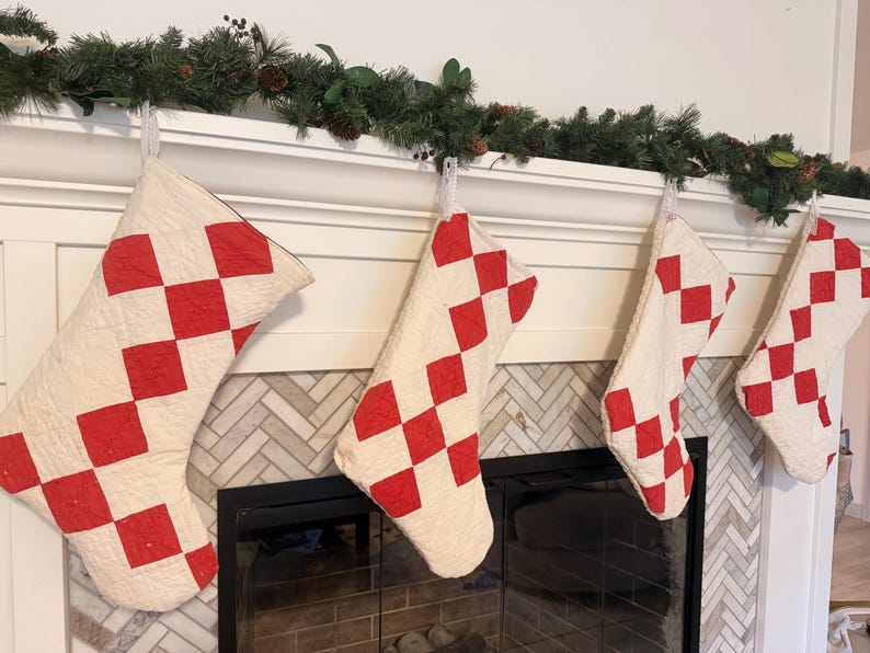 May include: Four quilted Christmas stockings featuring a repeating red and off-white diamond checkerboard pattern. Each stocking is constructed with visible stitching and hangs from a white mantelpiece, displaying a consistent geometric design.