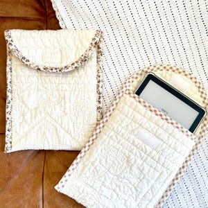 May include: Two quilted cream-colored e-reader cases. One case is closed, and the other is open, holding an e-reader. Both cases have a floral trim and a quilted pattern. The cases are on a brown surface.