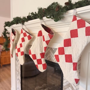 May include: Four quilted Christmas stockings hanging in a row, each featuring a red and white checkered pattern. The stockings are constructed with a visible stitched texture and have a red and black plaid fabric lining visible at the top opening.
