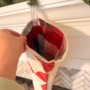 May include: A Christmas stocking featuring a quilted white exterior with red accents and a red, green, and white plaid interior lining. The stocking includes a white lace loop for hanging and is constructed from fabric with visible stitching.