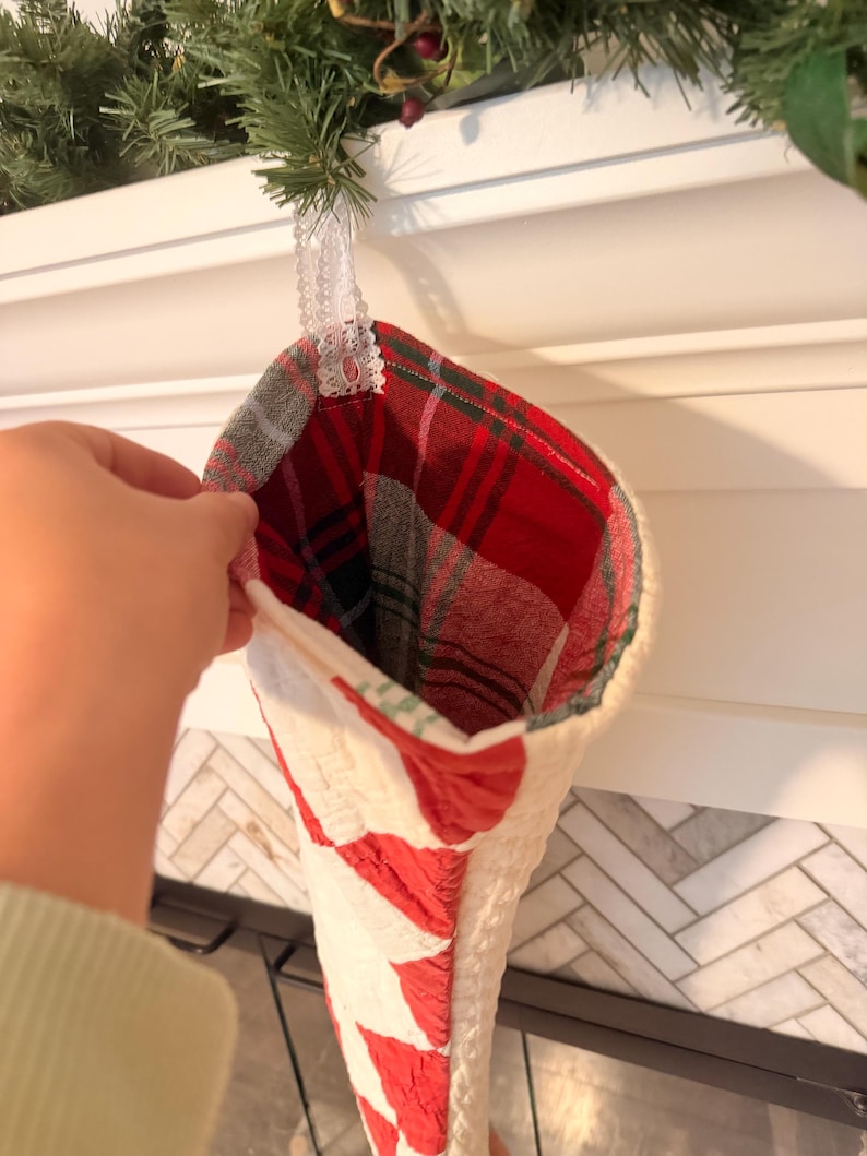 May include: A Christmas stocking featuring a quilted white exterior with red accents and a red, green, and white plaid interior lining. The stocking includes a white lace loop for hanging and is constructed from fabric with visible stitching.