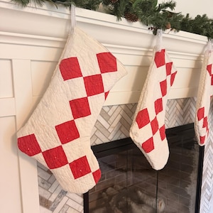 May include: A set of three quilted Christmas stockings featuring a white base with a red diamond-shaped checkerboard pattern. Each stocking is constructed with visible stitching and includes a white fabric loop for hanging on a fireplace mantel.