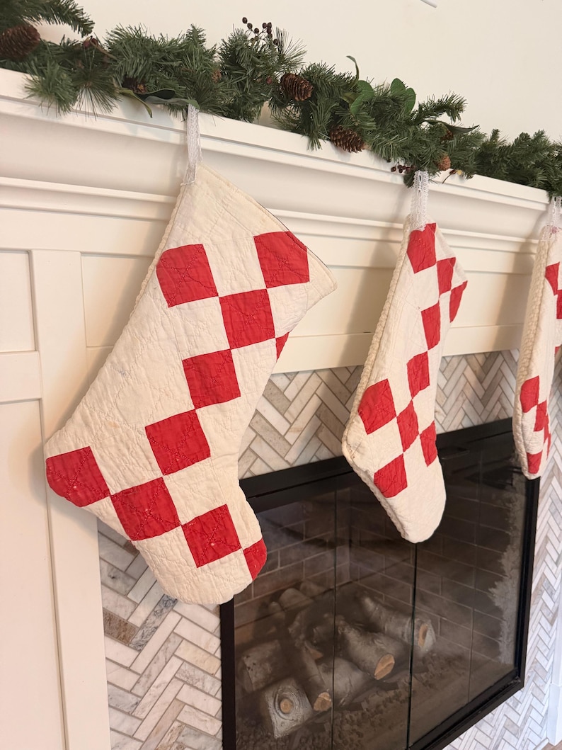 May include: A set of three quilted Christmas stockings featuring a white base with a red diamond-shaped checkerboard pattern. Each stocking is constructed with visible stitching and includes a white fabric loop for hanging on a fireplace mantel.