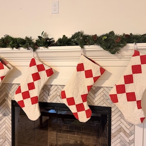 May include: Four quilted Christmas stockings hanging in a row, each featuring a repeating pattern of red diamond shapes on a cream-colored background. The stockings are constructed with visible stitching and have a soft, textured fabric appearance.