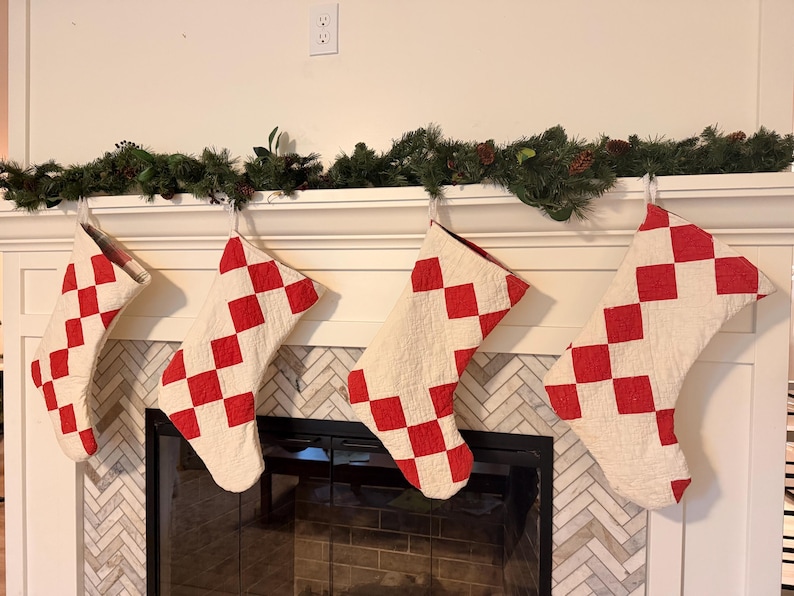 May include: Four quilted Christmas stockings hanging in a row, each featuring a repeating pattern of red diamond shapes on a cream-colored background. The stockings are constructed with visible stitching and have a soft, textured fabric appearance.