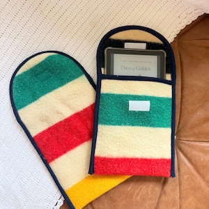 May include: Two rectangular felt sleeves with rounded tops, featuring horizontal stripes in green, cream, red, and yellow with dark blue trim. One sleeve contains an e-reader displaying the text Theo of Golden. Both sleeves have white velcro closures.