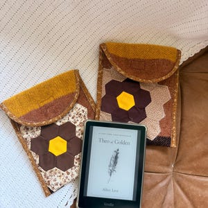 May include: Two fabric sleeves featuring a patchwork flower design with brown, yellow, and floral patterns, each with a mustard yellow flap. A black e-reader is positioned in front, displaying the book cover for Theo of Golden by Allen Levi.