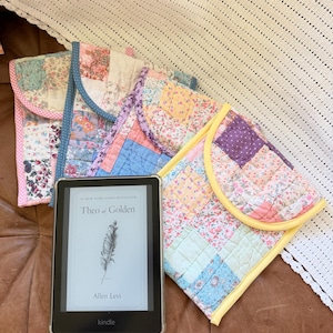 May include: A black Kindle e-reader displaying the book Theo of Golden by Allen Levi, positioned next to four quilted fabric sleeves. Each sleeve features a patchwork design of various floral and patterned fabrics with colorful fabric trim.