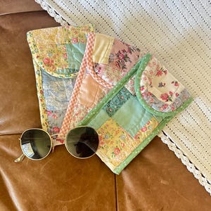 May include: Three quilted eyeglass cases in pastel colors with floral patterns and a pair of round sunglasses with gold frames. The cases are made of patchwork fabric and have a flap closure. The sunglasses are resting on a brown leather surface.