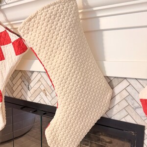 May include: A cream-colored quilted Christmas stocking with a textured, raised dot pattern. The stocking features a red fabric lining visible along the side edge and is suspended by a small white loop at the top, showcasing a classic boot shape.