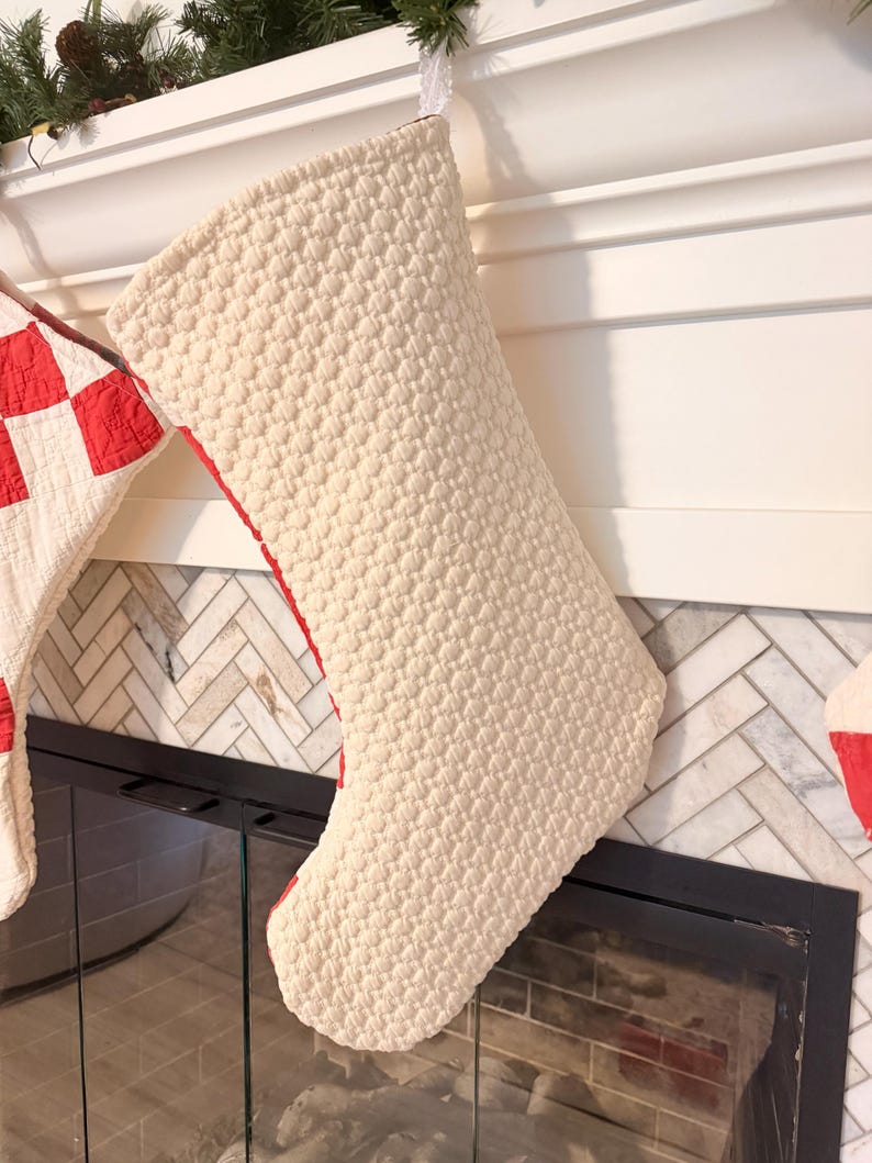 May include: A cream-colored quilted Christmas stocking with a textured, raised dot pattern. The stocking features a red fabric lining visible along the side edge and is suspended by a small white loop at the top, showcasing a classic boot shape.