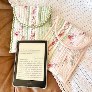 May include: Two quilted Kindle e-reader covers with floral and striped patterns. One cover has a green gingham flap, the other a pink trim. A Kindle e-reader displays text. The covers rest on a brown leather surface and a white crocheted blanket.