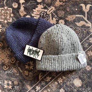 May include: Two knit beanies, one in dark navy blue and one in light grey with black flecks. The navy beanie features a rectangular white tag with black stylized text. The grey beanie has a white rectangular tag with the text The Winslow.
