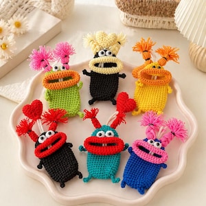 Puede incluir: Una colección de seis pequeños llaveros de monstruos tejidos a ganchillo de colores. Cada criatura presenta una boca ancha con dos dientes blancos, dos ojos redondos y dos pompones de hilo o adornos en forma de corazón. Los cuerpos son verde, negro, amarillo, turquesa y azul.