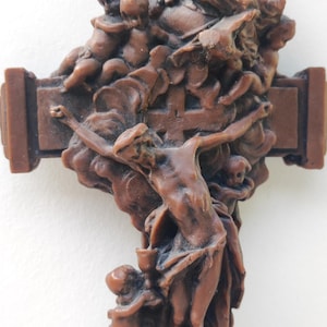 May include: A brown, molded resin wall crucifix featuring a detailed relief sculpture. The design depicts a central figure on a cross, surrounded by multiple smaller figures, including cherubs and a bearded figure at the top, all in a monochromatic finish.