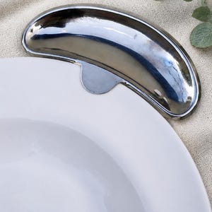 Peut inclure: Un accessoire de vaisselle en métal argenté poli en forme de croissant, conçu pour se fixer sur le bord d'une assiette en céramique blanche. L'élément incurvé présente une surface lisse et réfléchissante avec une petite languette de fixation.