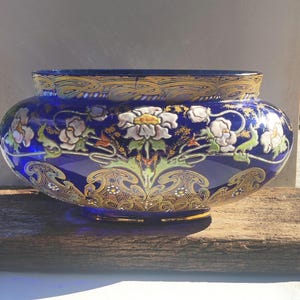 Op de afbeelding: Een kobaltblauwe glazen kom met een ingewikkeld handgeschilderd bloemmotief in wit, groen en geel, geaccentueerd met uitgebreide gouden filigraanpatronen rond de rand en de voet, wat een decoratief object met een brede, ronde vorm creëert.
