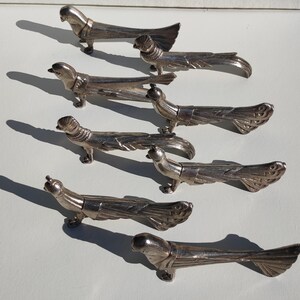Peut inclure: Un ensemble de six repose-couteaux en métal argenté en forme d'oiseau. Chaque pièce présente une forme aviaire stylisée avec une longue queue détaillée et de petites pattes. Les figurines sont disposées en quinconce, révélant leur fini métallique.