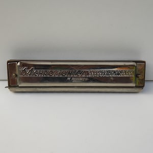 Puede incluir: Una armónica M. Hohner 64 Chromonica modelo profesional de tono plateado con acabado metálico. La placa frontal presenta un texto grabado que dice The 64 Chromonica, Professional Model y M. Hohner Germany, asegurada con tornillos.