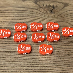 May include: A collection of nine small, orange, oval-shaped game tokens. Each token features white printed text displaying a plus one over plus one fraction, followed by two-digit numbers including 02, 05, 10, 00, 55, and 22, with white gear-like edges.
