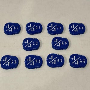 May include: Ten small, oval-shaped blue game tokens arranged in three rows. Each token features a white printed graphic of a plus one over plus one fraction symbol, accompanied by various small numerical digits printed to the right of the fraction.
