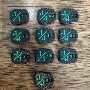 May include: Ten black, textured, rounded tokens featuring a bright green printed graphic of a plus one over plus one symbol and two-digit numbers ranging from 11, 22, 33, 44, 55, 66, 77, 88, 99, to 00, arranged in a grid pattern on a wooden surface.