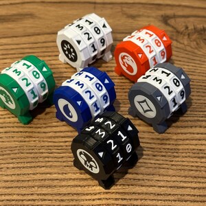 May include: A collection of six cylindrical life counters in white, orange, green, blue, gray, and black. Each counter features rotating number wheels with digits zero through nineteen and a unique icon on the side, including a skull, tree, and flame.