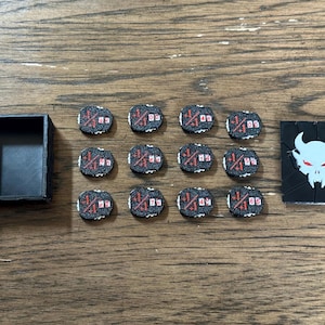 May include: A black rectangular storage box, twelve small circular black tokens with red and white numerical markings, and a black rectangular card featuring a white horned skull graphic with red eyes, all arranged on a wooden surface.