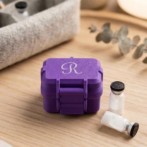 May include: A small, rectangular, purple storage case with a glittery finish and the letter R engraved on the lid. The case features a hinged latch closure. Two small glass vials with black caps and white powder contents are placed beside the case.