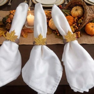 May include: Three white fabric napkins held by gold-colored metal napkin rings shaped like maple leaves. The napkins are arranged upright on a dark wooden table, each featuring a detailed leaf design on the ring that secures the folded cloth.
