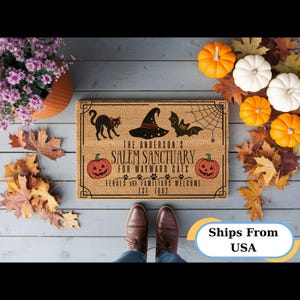 Peut inclure: Un paillasson rectangulaire en fibre de coco beige avec des illustrations noires d'un chat, d'un chapeau de sorcière, d'une chauve-souris, d'une toile d'araignée et deux citrouilles orange. Le texte indique: THE ANDERSON'S SALEM SANCTUARY FOR WAYWARD CATS, FERALS AND FAMILIARS WELCOME, EST. 1692.
