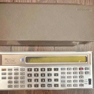 May include: A vintage Sharp EL-5100 scientific calculator with a silver-toned faceplate, a yellow liquid crystal display, and a full QWERTY-style keyboard layout, accompanied by a matching brown plastic protective slide-on cover case.
