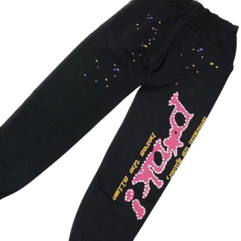 K&ouml;nnte beinhalten: Schwarze Jogginghose mit einer rosa gepunkteten Grafik mit der Aufschrift "paki" und gelbem Text mit "heres the slime" und "never mind the spider". Der Stoff ist mit kleinen, verstreuten mehrfarbigen Sternen auf den Oberschenkeln verziert.