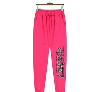 K&ouml;nnte beinhalten: Knallpinke Jogginghose mit elastischem Bund und Kordelzug, B&uuml;ndchen an den Kn&ouml;cheln und verstreuten, bunten Farbspritzern. Auf dem linken Bein befindet sich eine gro&szlig;e, schwarze, mit Nieten besetzte Grafik mit dem Text "SP5DER" und Text.
