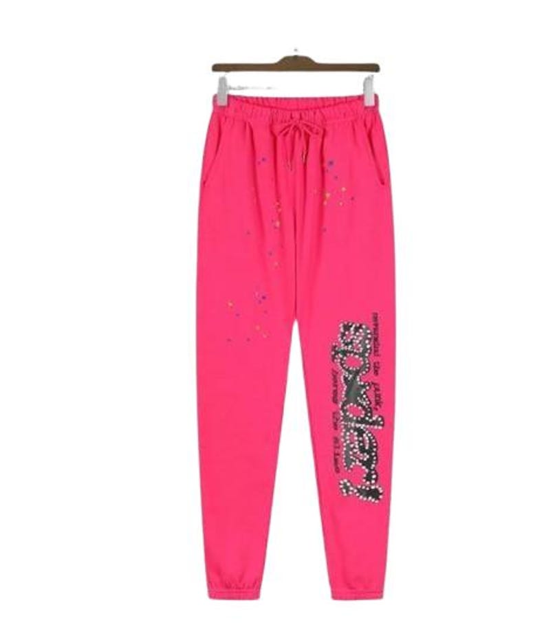 K&ouml;nnte beinhalten: Knallpinke Jogginghose mit elastischem Bund und Kordelzug, B&uuml;ndchen an den Kn&ouml;cheln und verstreuten, bunten Farbspritzern. Auf dem linken Bein befindet sich eine gro&szlig;e, schwarze, mit Nieten besetzte Grafik mit dem Text "SP5DER" und Text.