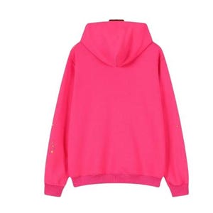 K&ouml;nnte beinhalten: Ein leuchtend neonpinker Kapuzenpullover mit langen &Auml;rmeln, gerippten B&uuml;ndchen und einem passenden gerippten Saum. Die R&uuml;ckseite des Kleidungsst&uuml;cks zeigt ein kleines, verstreutes Muster aus winzigen gelben Sternen entlang der &Auml;rmelau&szlig;enseiten.