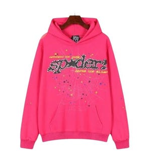 K&ouml;nnte beinhalten: Ein leuchtend pinkfarbener Kapuzenpullover mit einer gro&szlig;en schwarzen Grafik, die den Text "spider!", "demanded the punk" und "bares the slime" in Gelb zeigt. Das Design enth&auml;lt ein Spinnennetz-Muster mit kleinen bunten Sternen auf der Vorderseite.