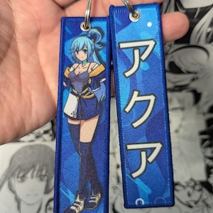 May include: Two rectangular fabric keychains with blue borders and metal rings. One features a blue-haired anime character in a blue and white outfit. The other displays the Japanese text "アクア" in white letters against a blue patterned background.